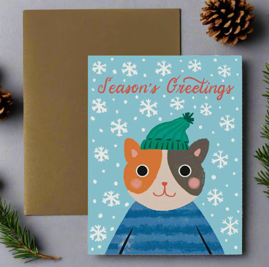Greeting card with a cat illustration wearing a green hat and blue sweater on a snowy background, with a brown envelope.